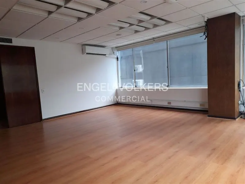Empty commercial space with wood floors, white walls, and large windows. A dark wood door is on the left, and an AC unit is above the Engel & Volkers logo.