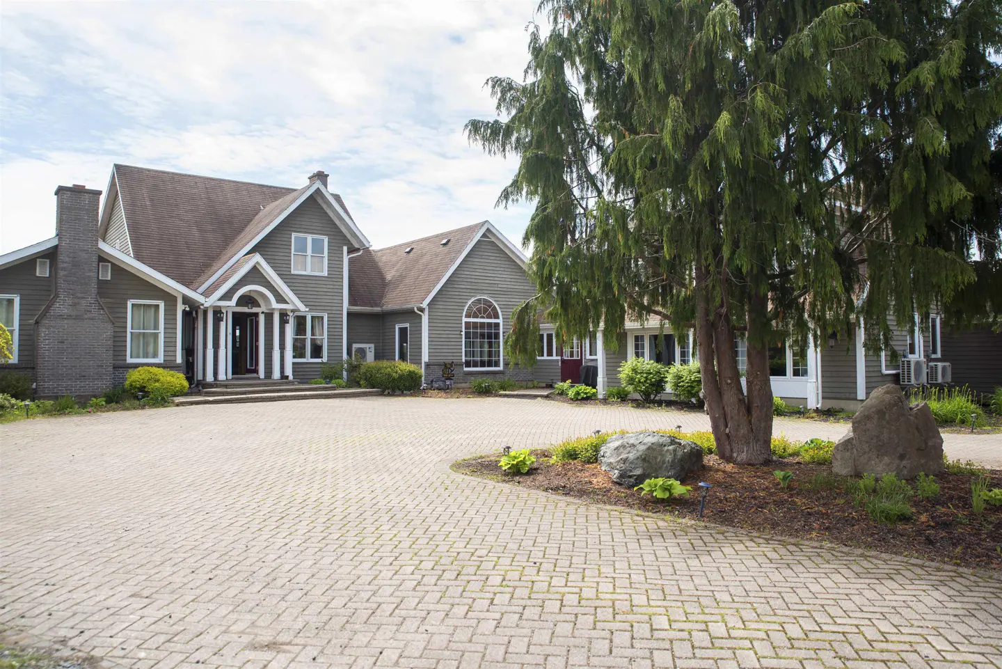 Exterior view of a large, two-story house with a brick driveway and a large tree in the front yard. The house is painted in a neutral color with white trim.