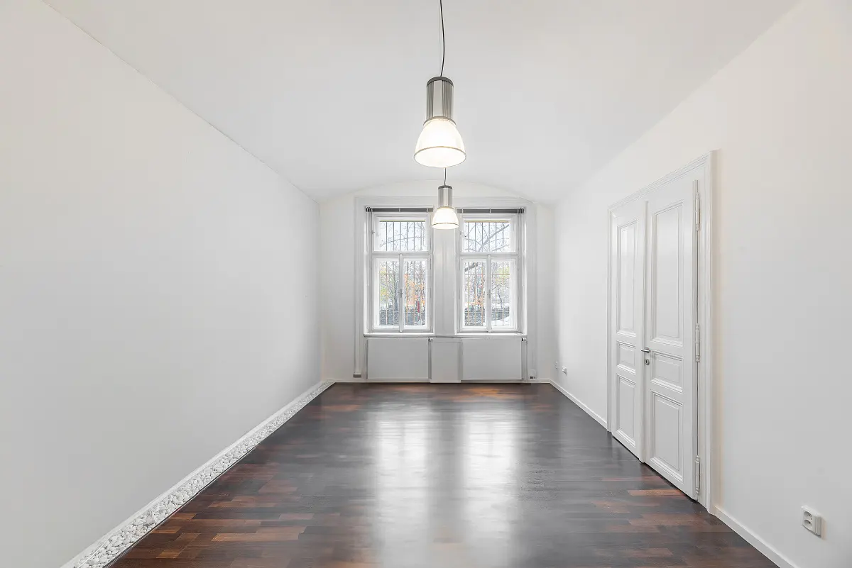 Empty room with dark wood floors, white walls, and two windows. Two pendant lights hang from the ceiling. A white door is on the right.