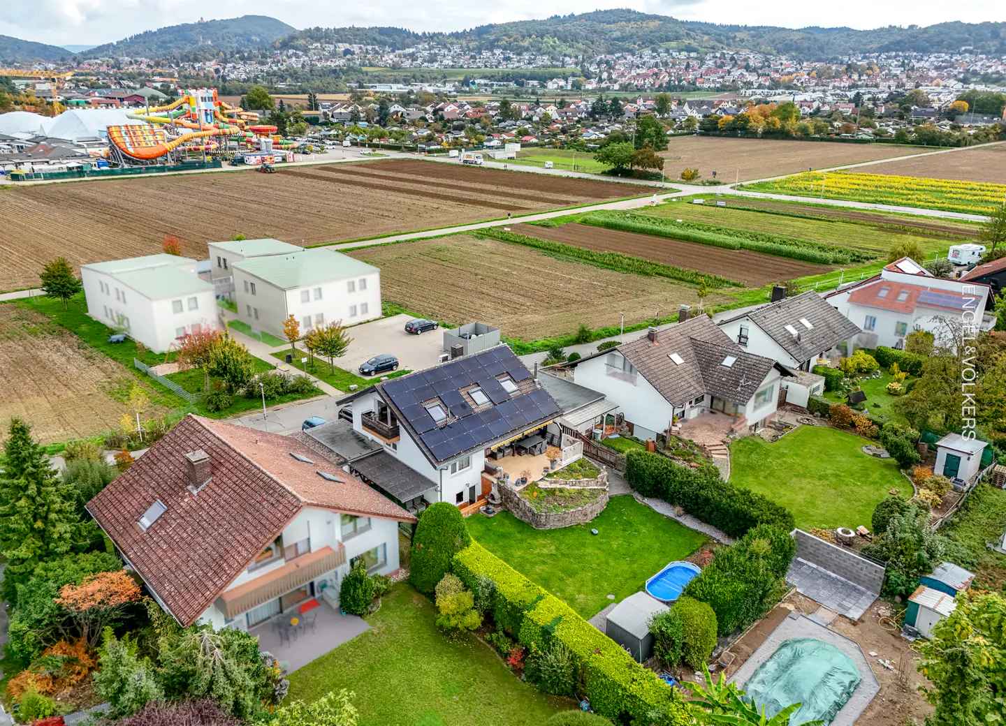 Aerial view of houses with green lawns, a water park in the background, and fields under a cloudy sky.