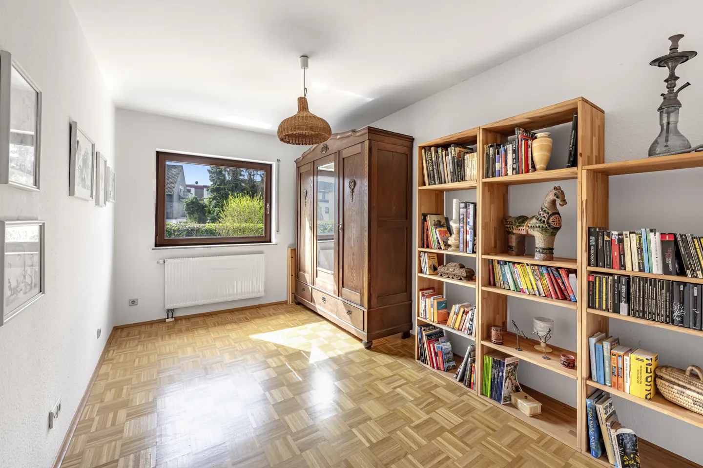 Bright room with parquet floor, white walls, window, radiator, wardrobe, and wooden bookshelves filled with books and decorative objects.