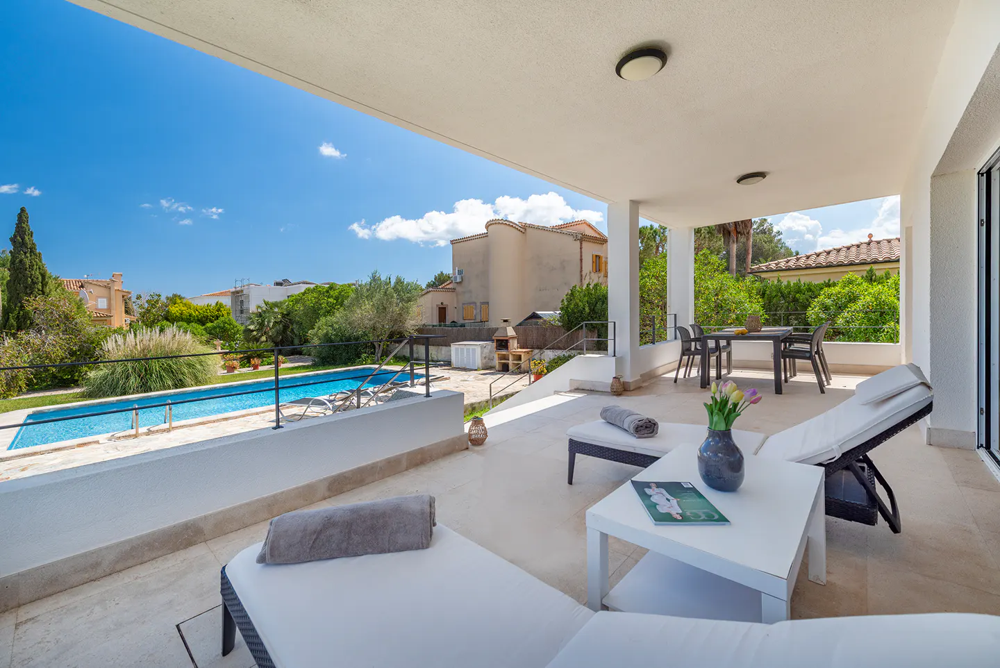 Covered patio with white lounge chairs, table with flowers, and a pool in the background on a sunny day.