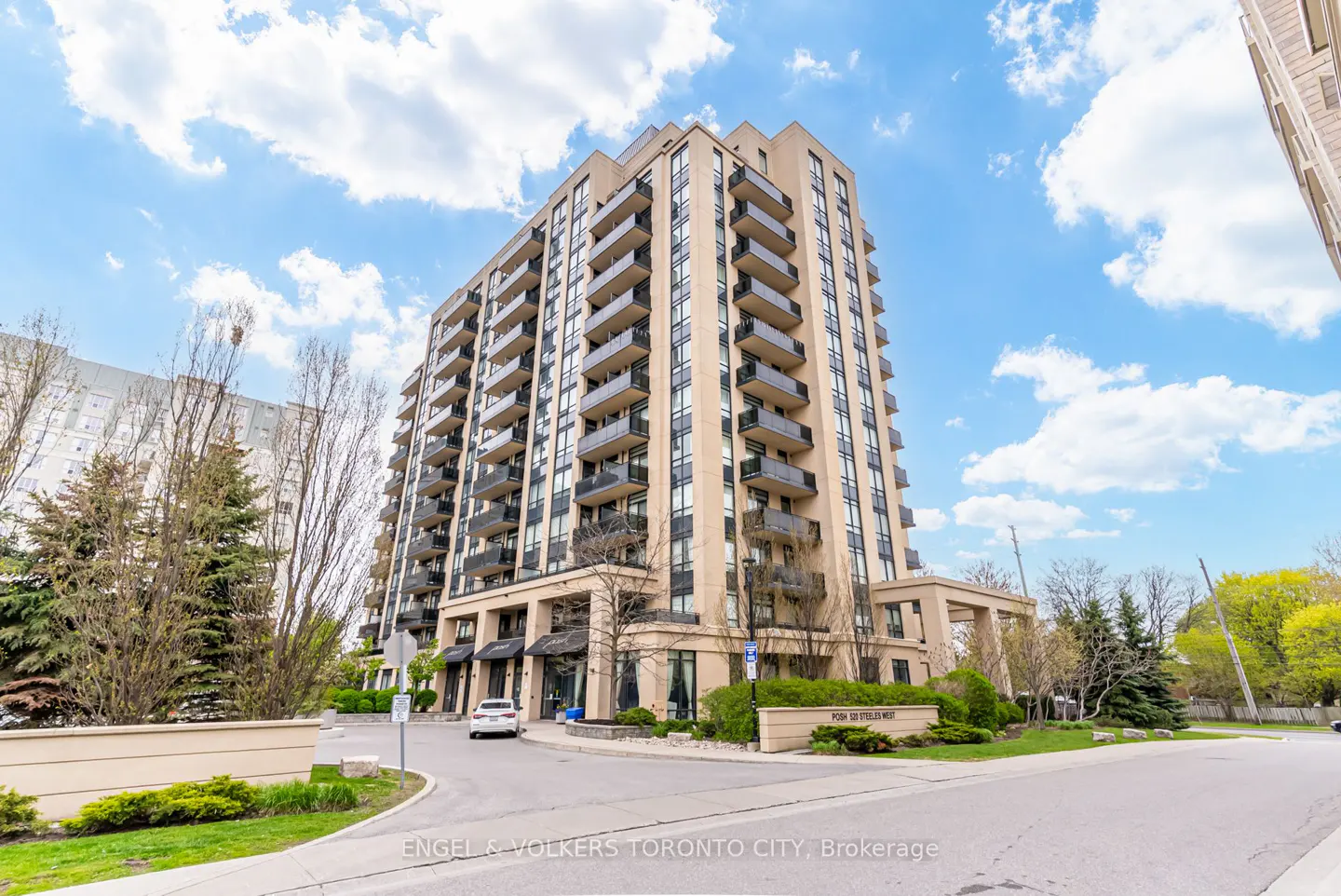 Exterior view of a tall, beige condo building with balconies under a blue sky with white clouds. A car is parked near the entrance.