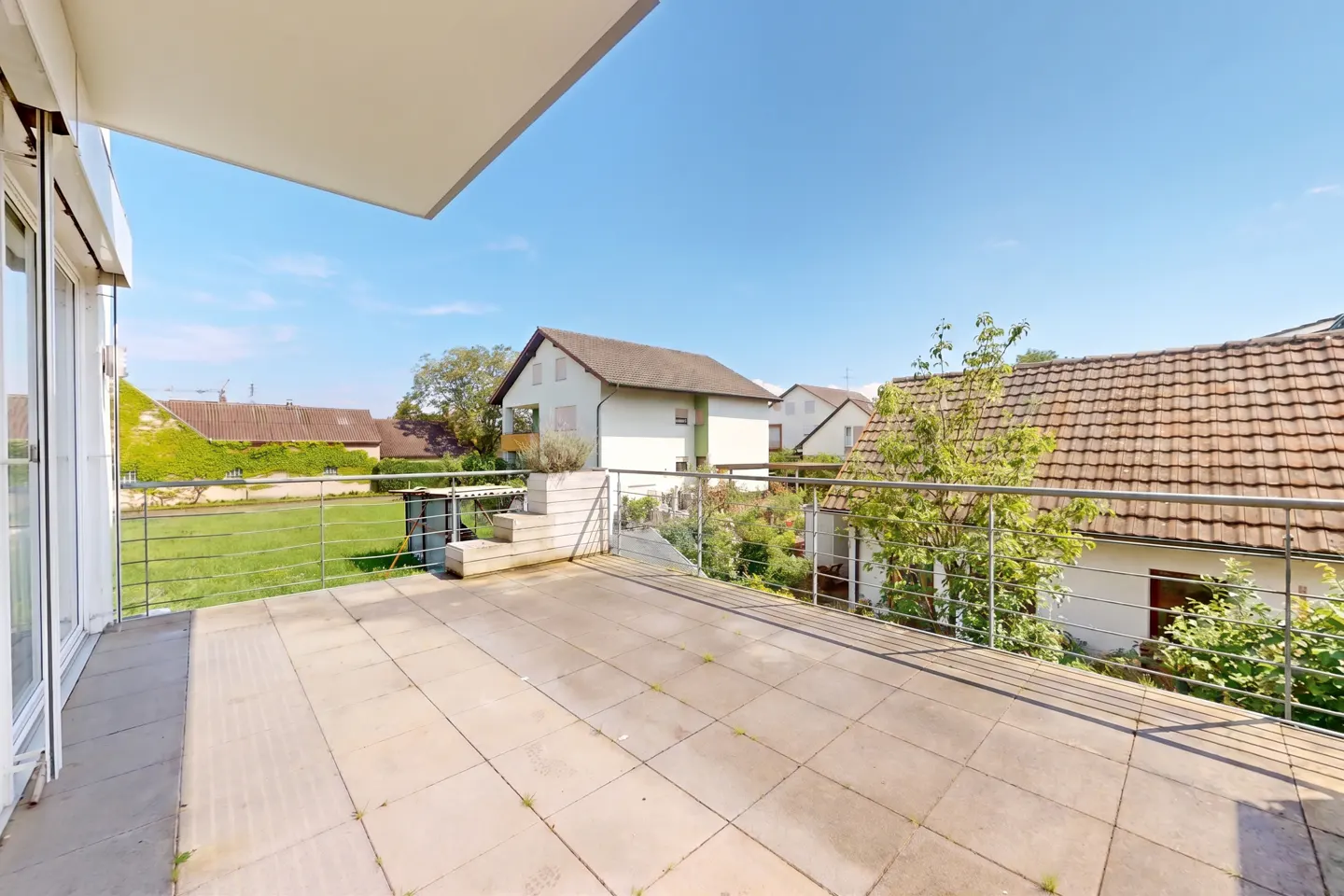A sunny balcony with beige tiles and a metal railing overlooks houses and green lawns.
