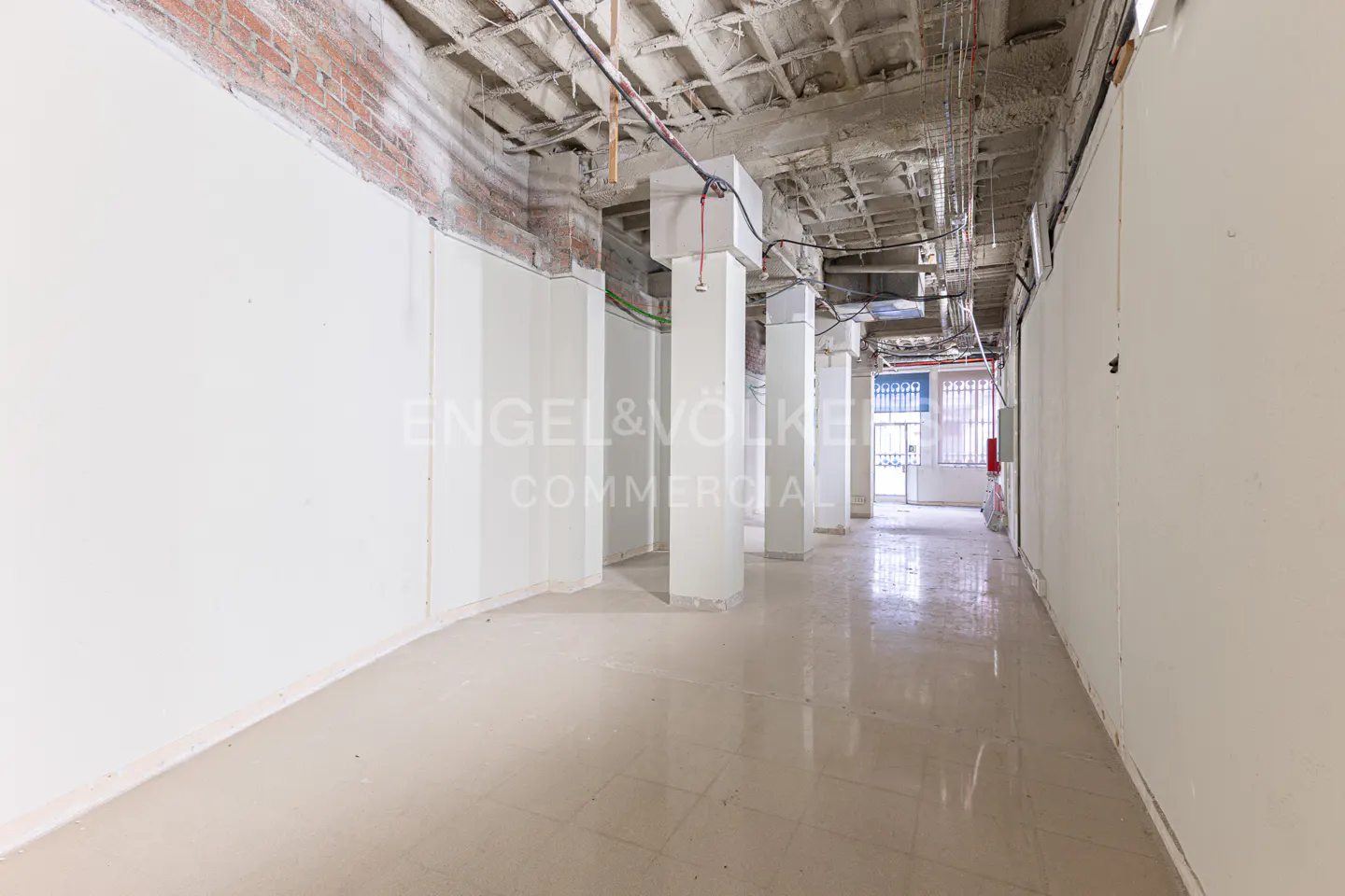Empty commercial space with white walls, columns, and a concrete ceiling with exposed wiring. The floor is light beige tile. "Engel & Völkers Commercial" is visible.