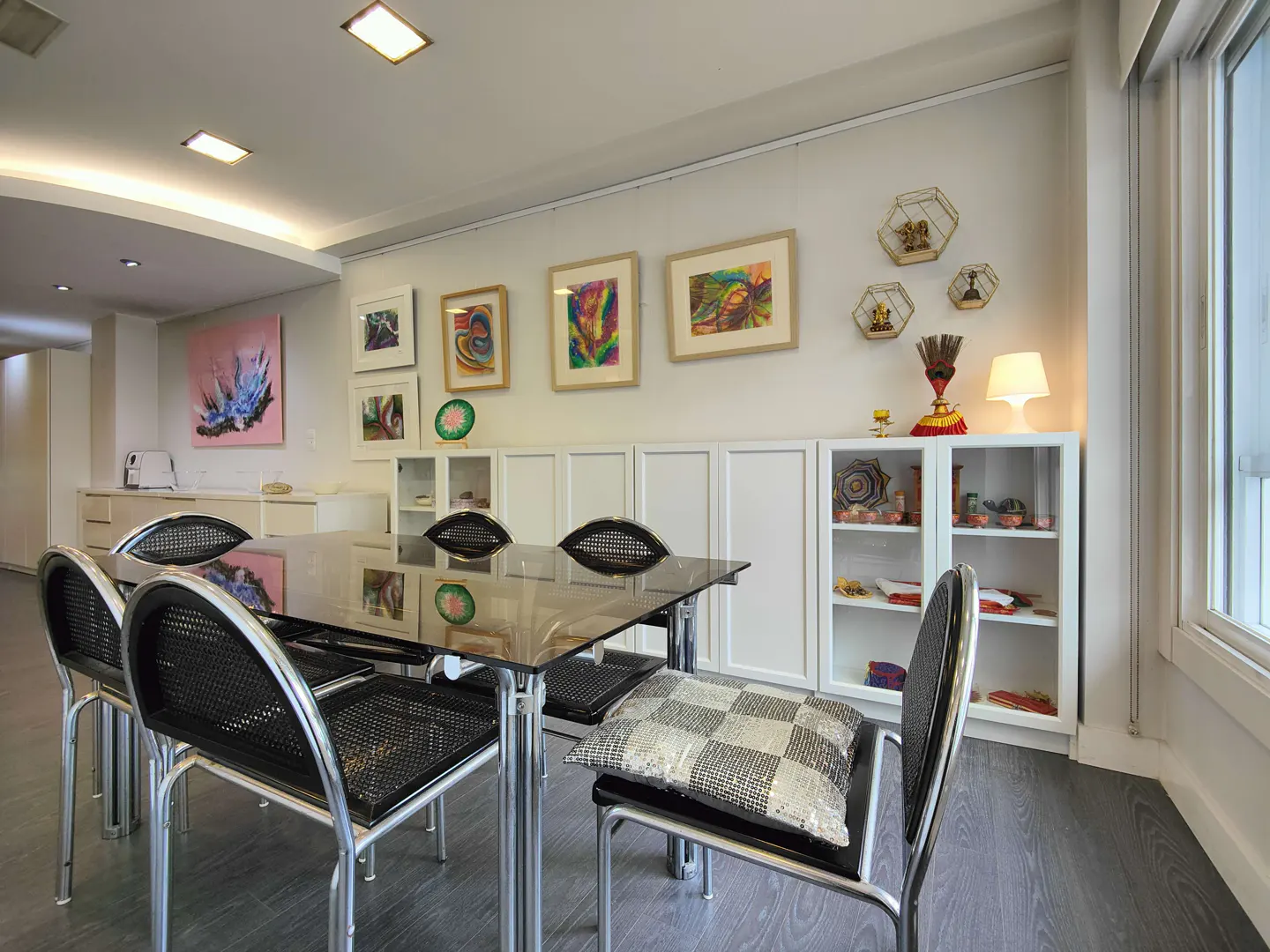 Bright dining room with a glass table, black chairs, and art on the white walls. A white cabinet displays decorative items.