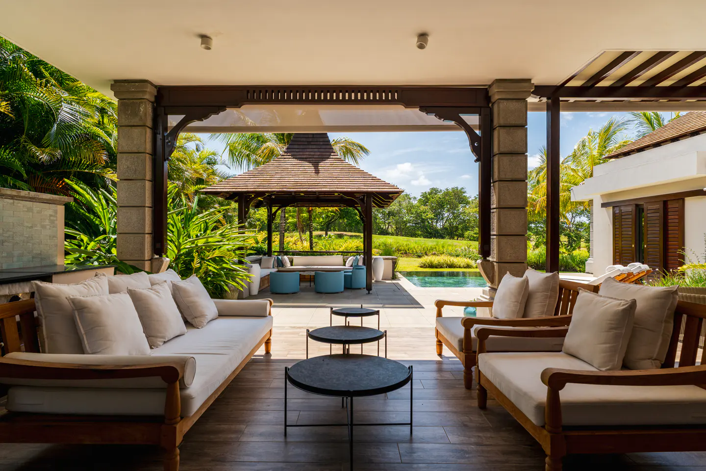 Covered patio with wood furniture and white cushions overlooks a gazebo, pool, and lush green landscape.