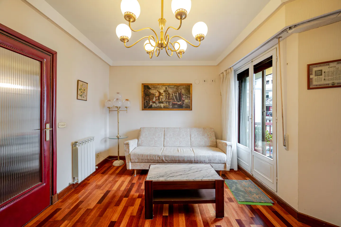 Living room with a beige sofa, marble-topped coffee table, chandelier, and a red door on a hardwood floor.