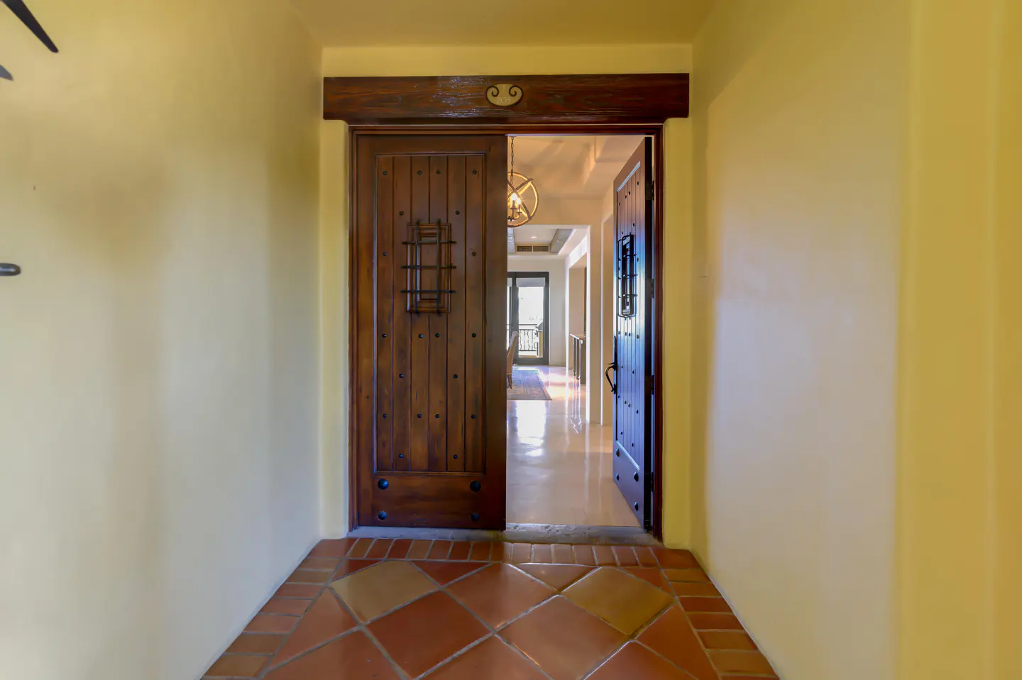 Open wooden double doors with iron accents lead to a bright hallway with tile flooring. Walls are painted a soft yellow.
