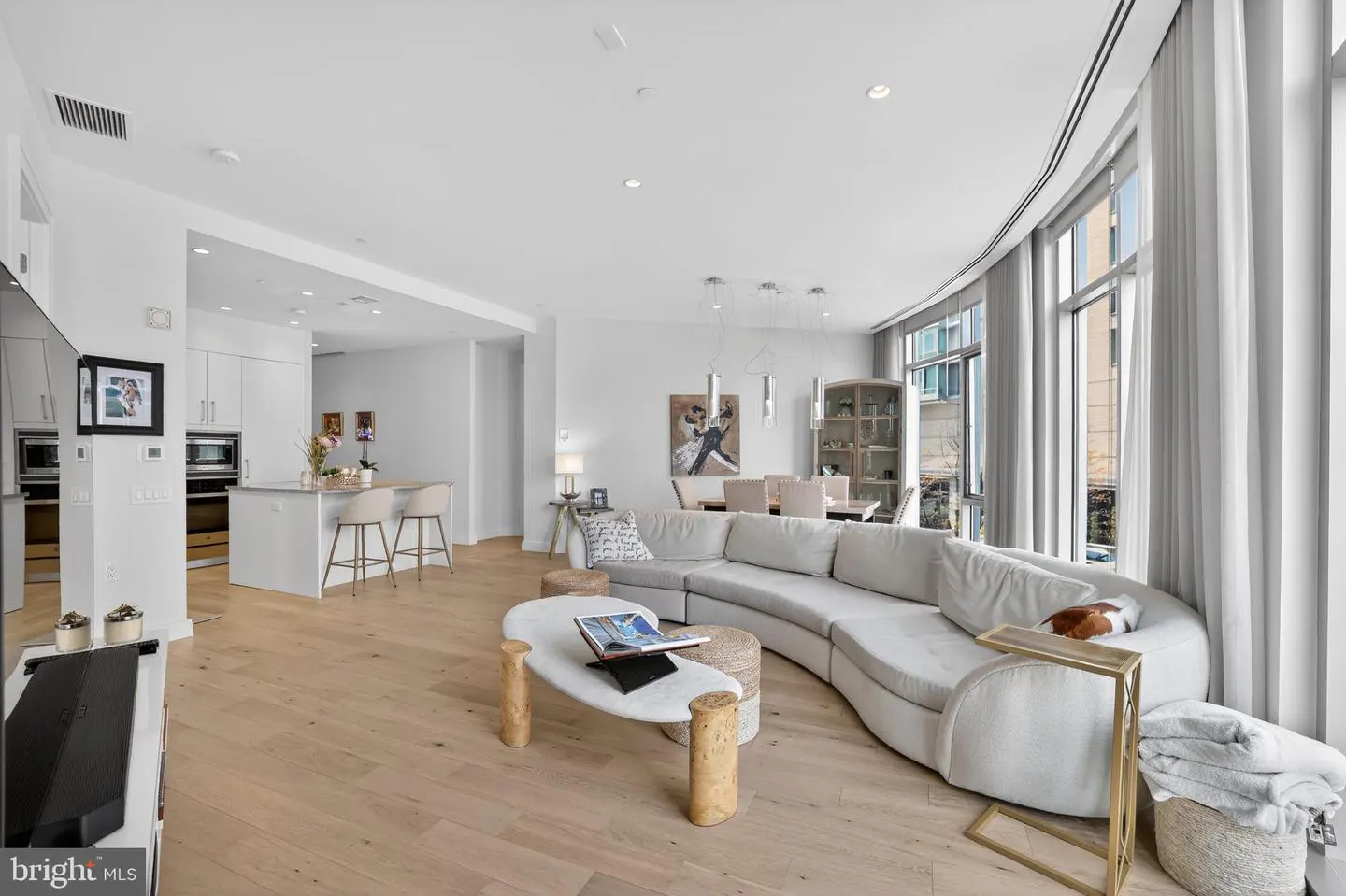 Bright, open-concept living space with a curved white sofa, marble coffee table, and light wood floors. Kitchen and dining area visible in the background.
