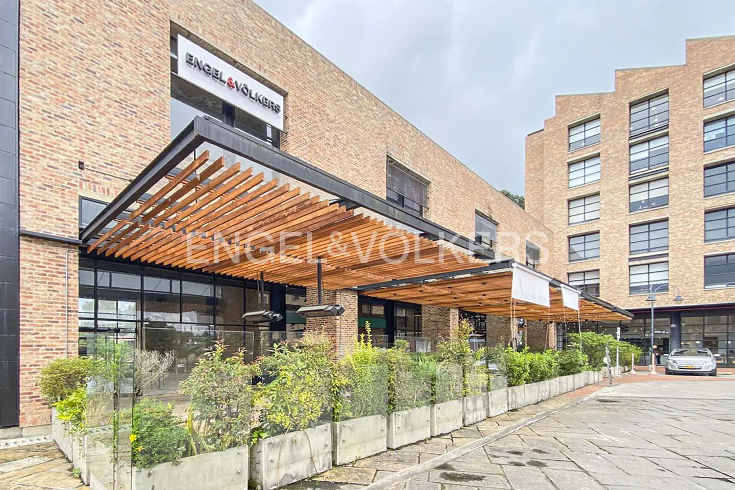 Exterior view of an Engel & Volkers office in a brick building with a wooden awning and greenery.
