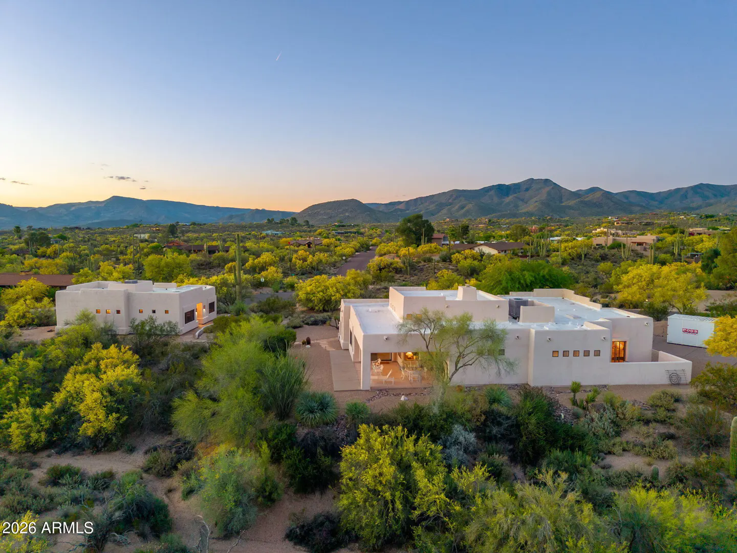 Aerial view of a modern, white, flat-roofed home surrounded by desert vegetation and mountains at dusk.