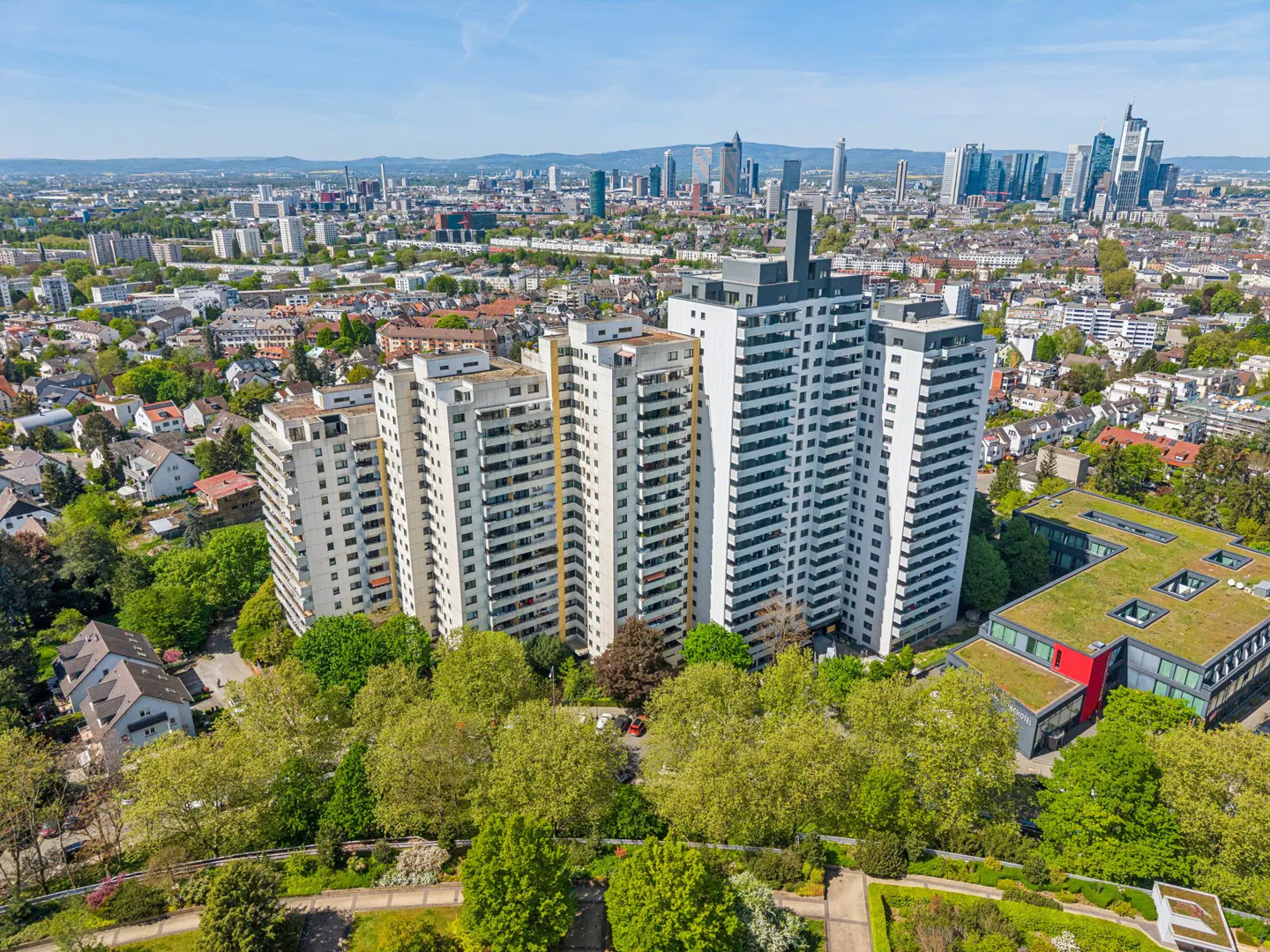 Aerial view of white apartment buildings surrounded by green trees, with a city skyline in the background under a blue sky.
