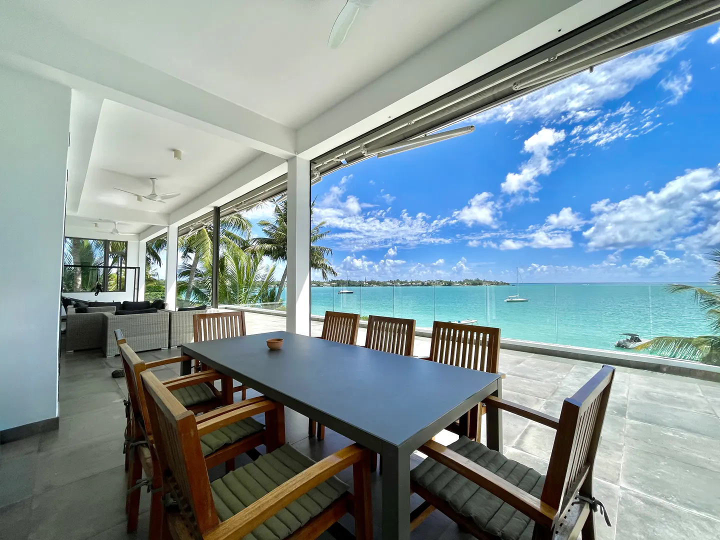Outdoor dining area with a gray table and wooden chairs overlooking a turquoise ocean and blue sky with clouds.