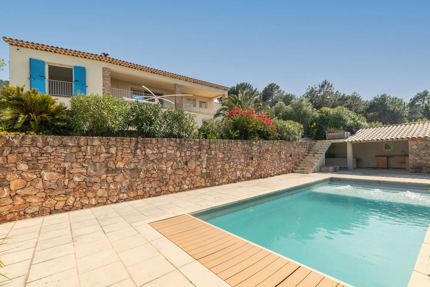 Exterior view of a two-story house with a pool, stone wall, and blue shutters on a sunny day.