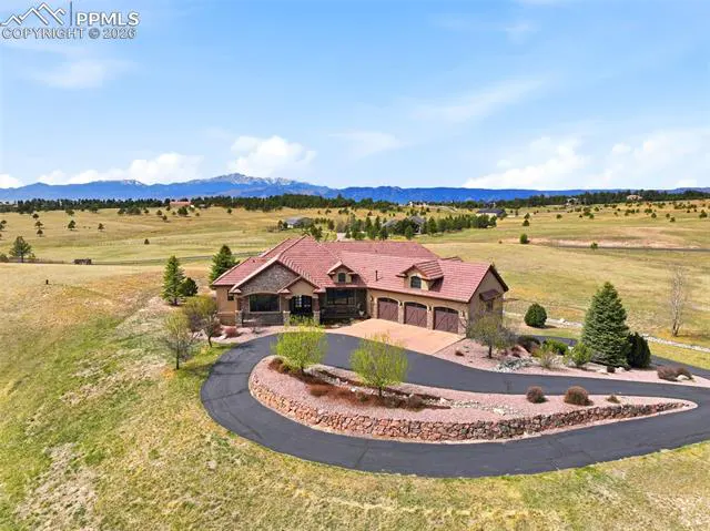 A tan house with a red tile roof and a three-car garage sits on a grassy lot with mountains in the background.
