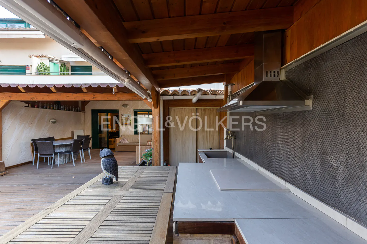 Outdoor kitchen with wood beams, gray backsplash, and white countertops. A wooden table with an owl statue sits nearby.