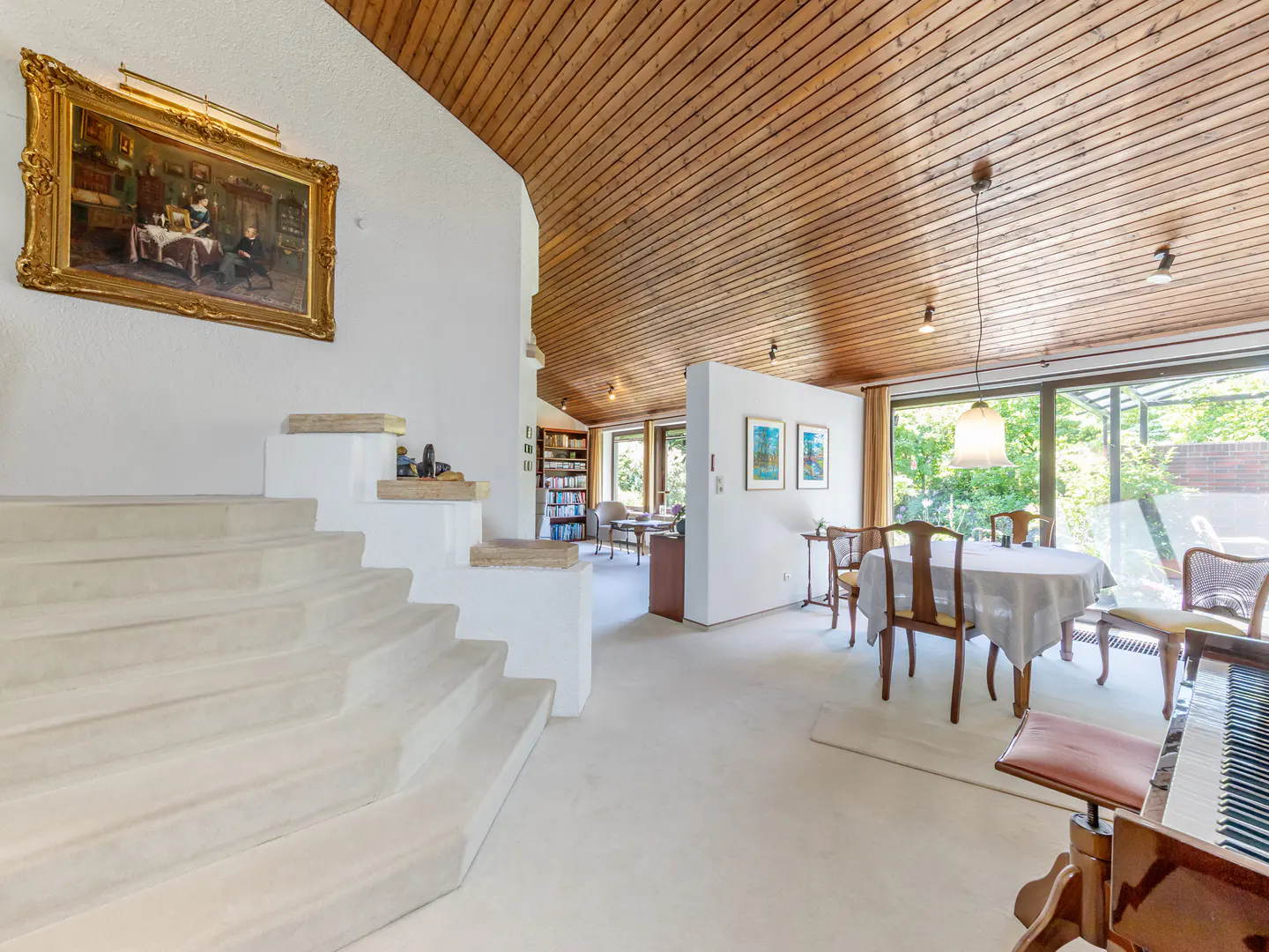 Spacious living area with white stairs, wood-paneled vaulted ceiling, dining set, and a piano under a large framed painting.