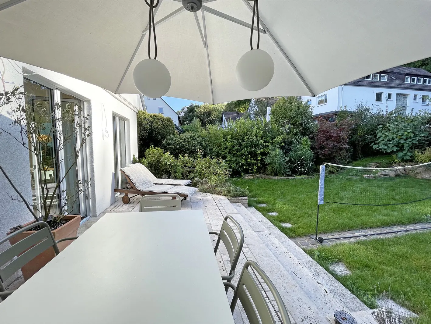 Outdoor patio with white table, chairs, and lounge chairs under a large umbrella. Green lawn and trees in the background.