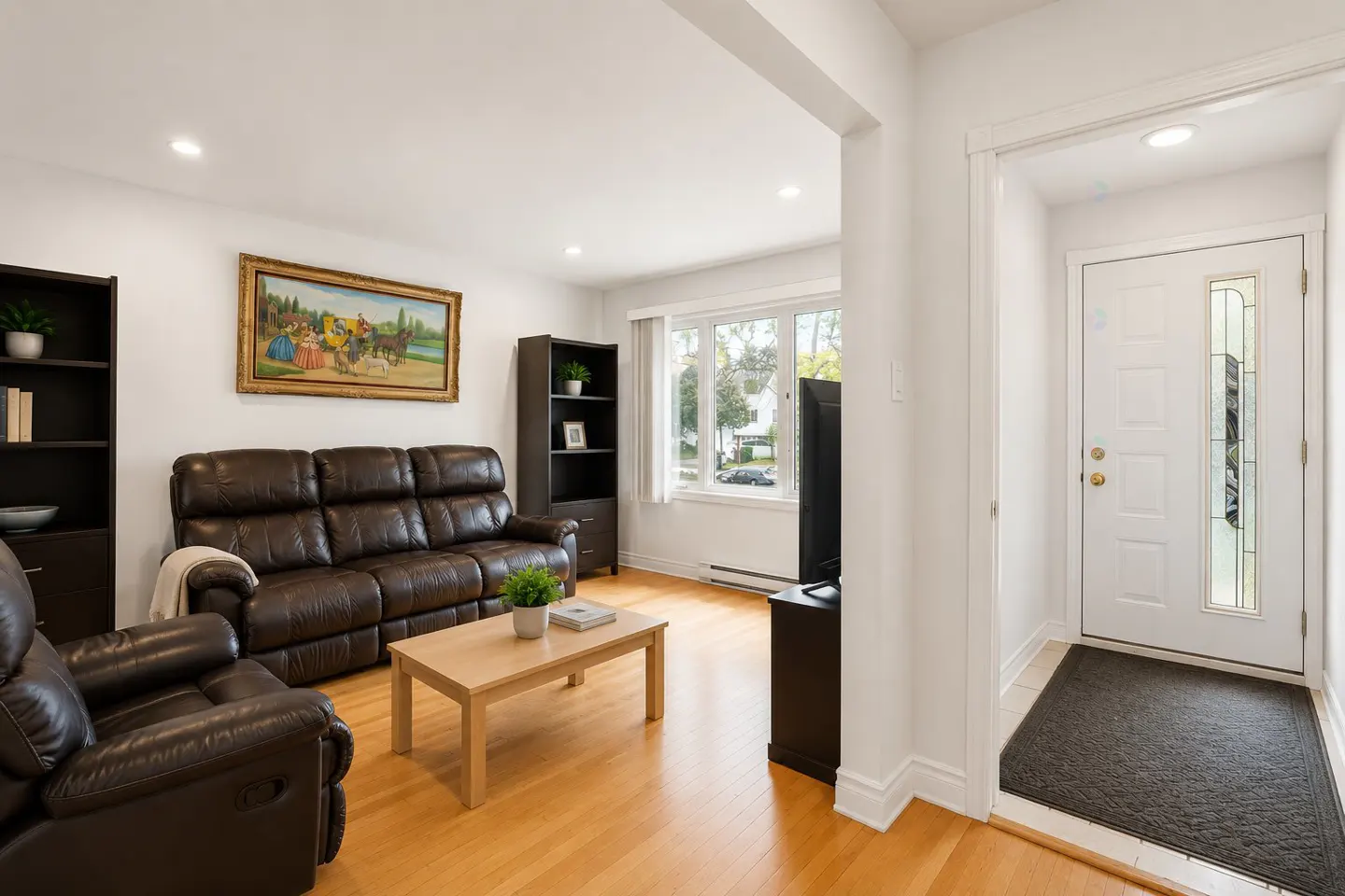 Bright living room with a brown leather sofa, wood floors, and a white front door. A painting hangs above the sofa.