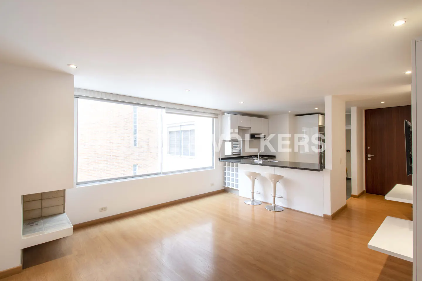 Bright, modern apartment with wood floors, white walls, and a large window. A kitchen island with bar stools separates the living area.