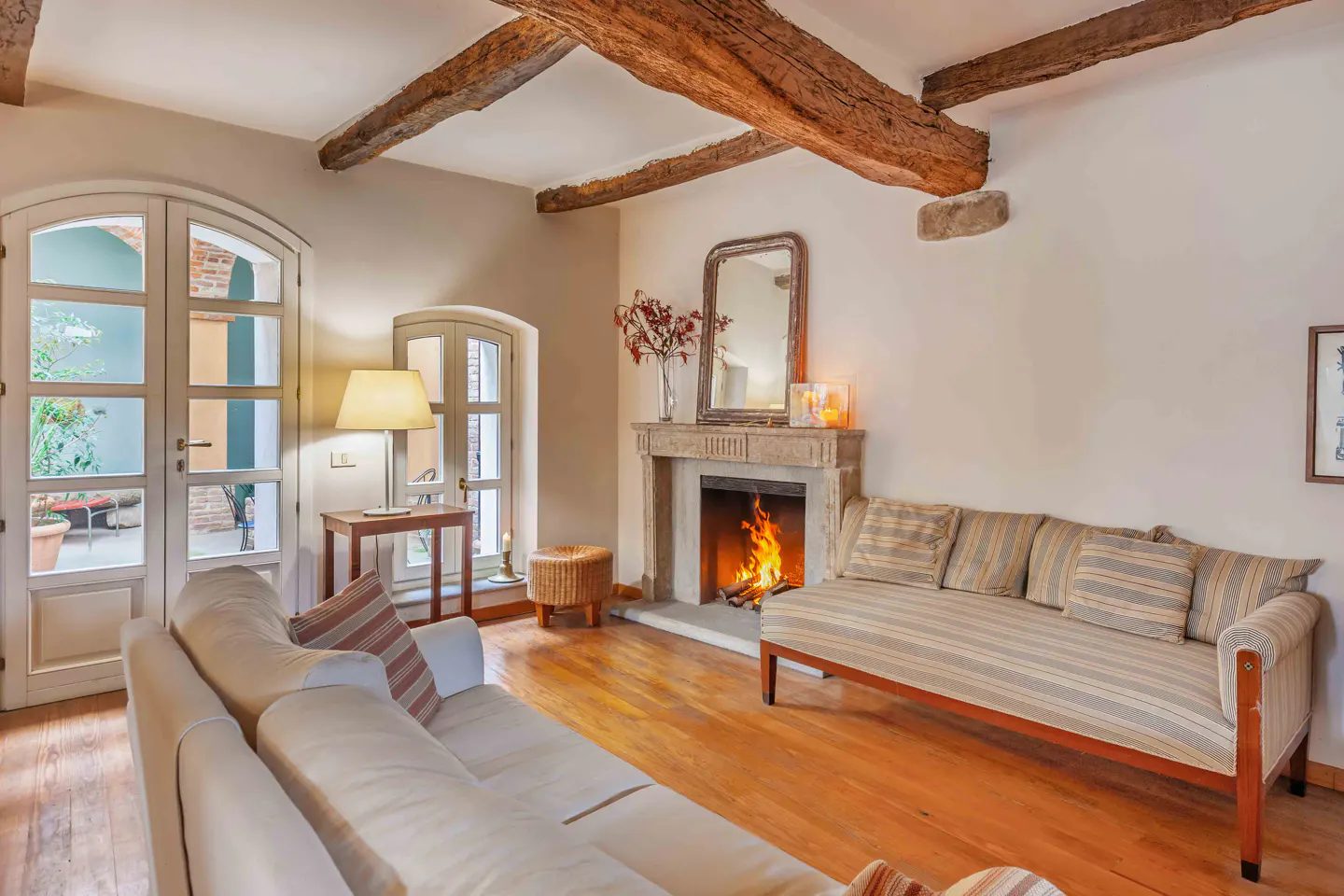 Living room with exposed wood beams, hardwood floors, and a stone fireplace with a fire burning. White sofa and striped settee.