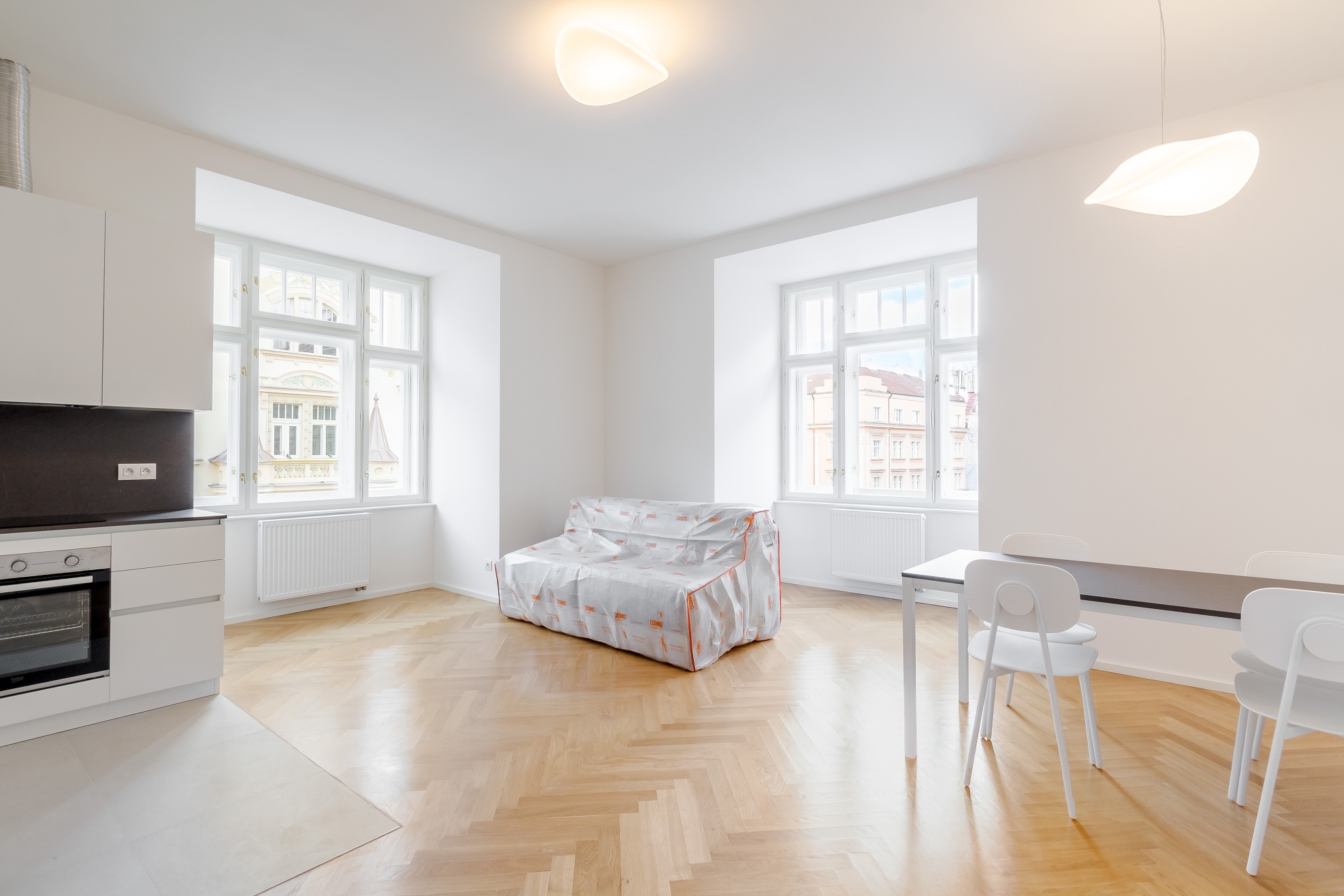 Bright, modern studio apartment with white kitchen, sofa covered in plastic, dining area, and light wood herringbone floors.