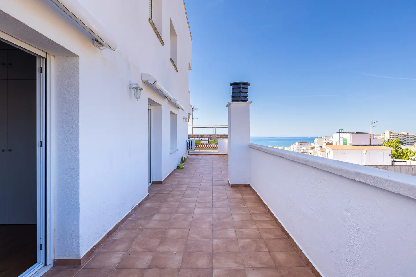 A bright, sunny balcony with brown tile flooring, white walls, and a view of the ocean and city skyline.