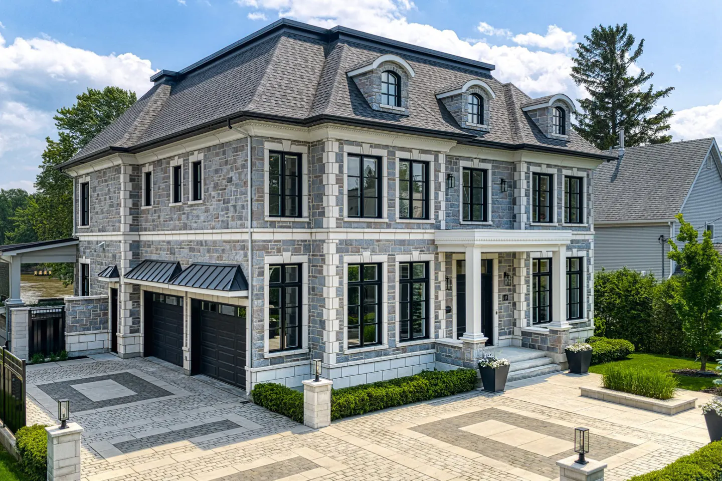 Exterior of a large, gray stone house with black framed windows and a patterned stone driveway.