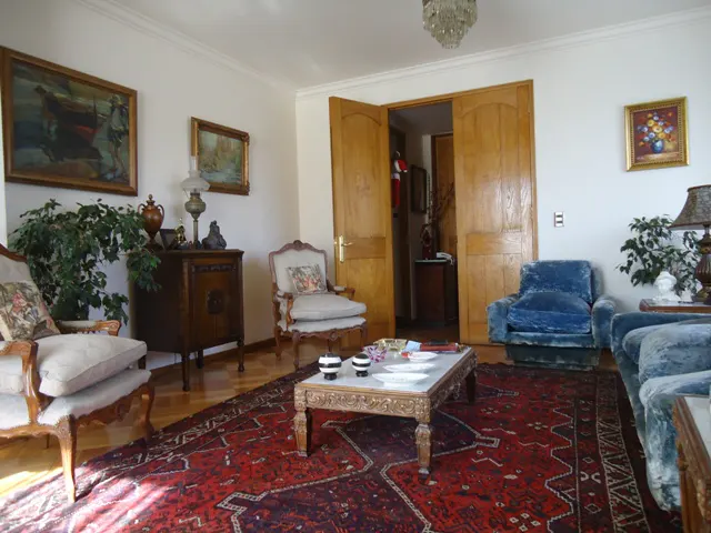 Living room with antique furniture, paintings, and a red patterned rug. A chandelier hangs from the ceiling. Double wooden doors are open in the background.