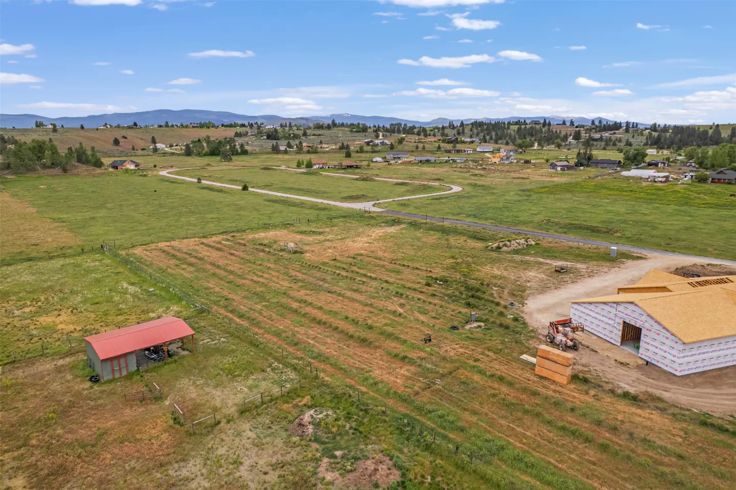 Aerial view of a rural property with a barn under construction, a red-roofed shed, and fields, under a blue sky with scattered clouds.