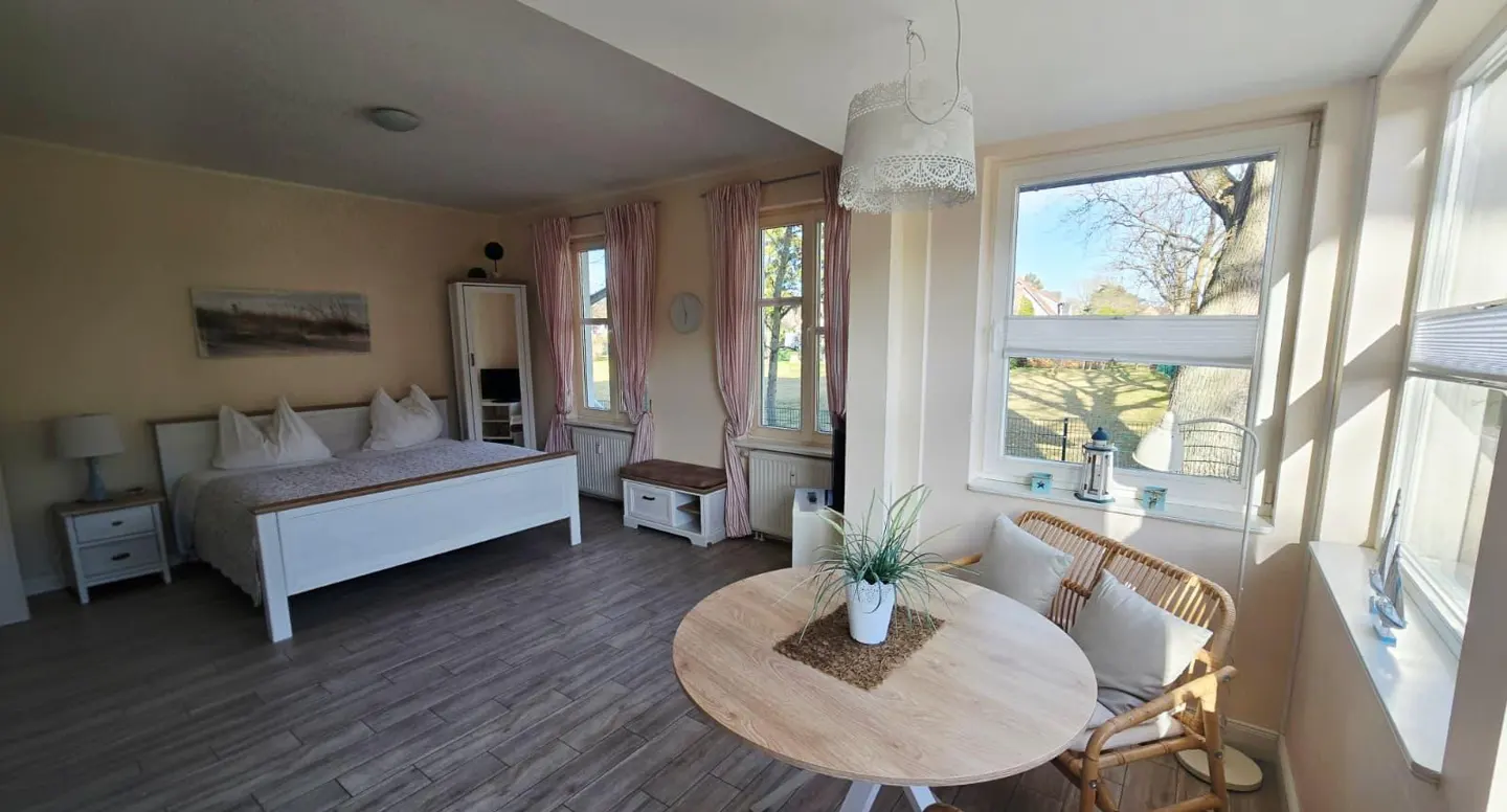 Bright studio apartment with a white bed, round table, and large windows overlooking a green lawn.