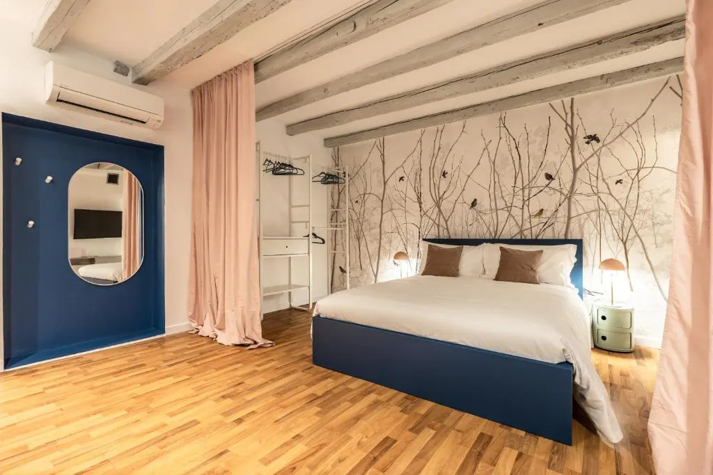 A bedroom with a blue bed frame, white bedding, and a tree-themed wallpaper. There is a blue door with a mirror and pink curtains.