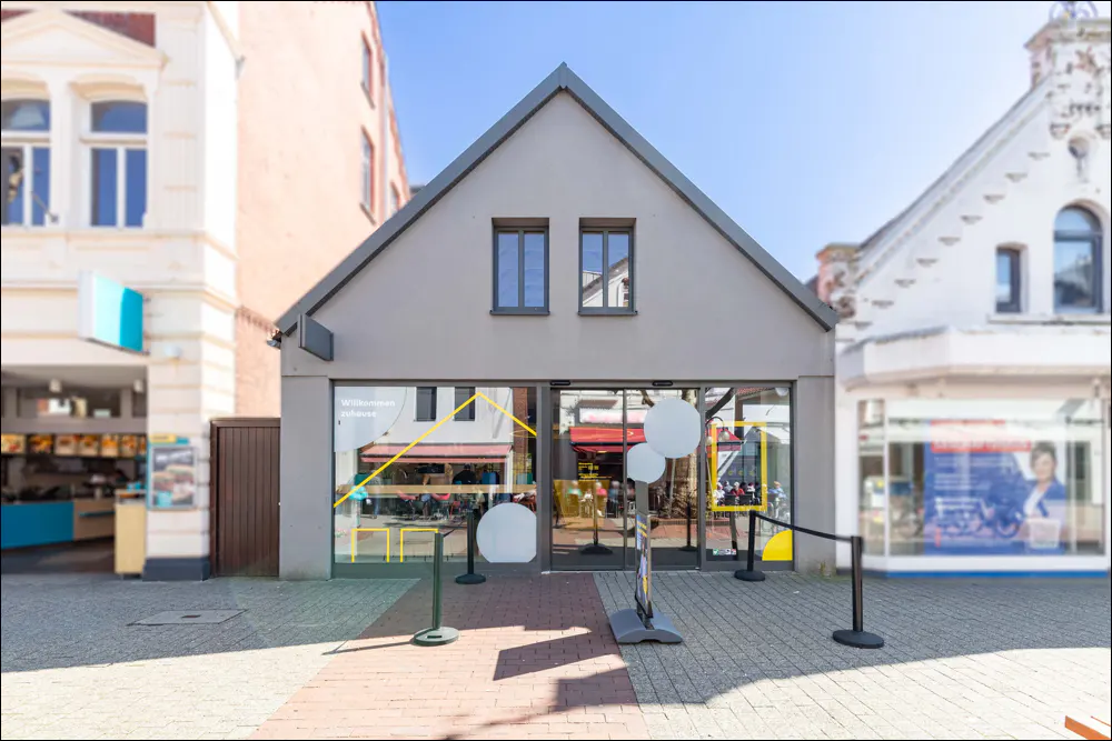 A modern grey building with a triangular roof and large glass storefront, situated between two traditional town buildings.