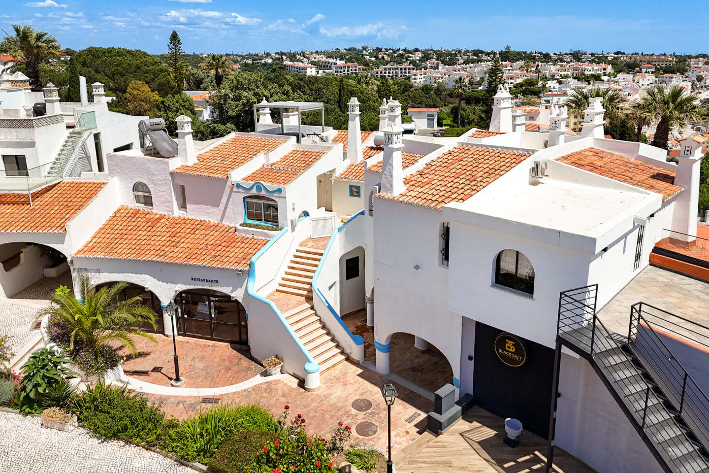 Aerial view of white buildings with orange tile roofs, blue trim, and lush greenery under a bright blue sky.