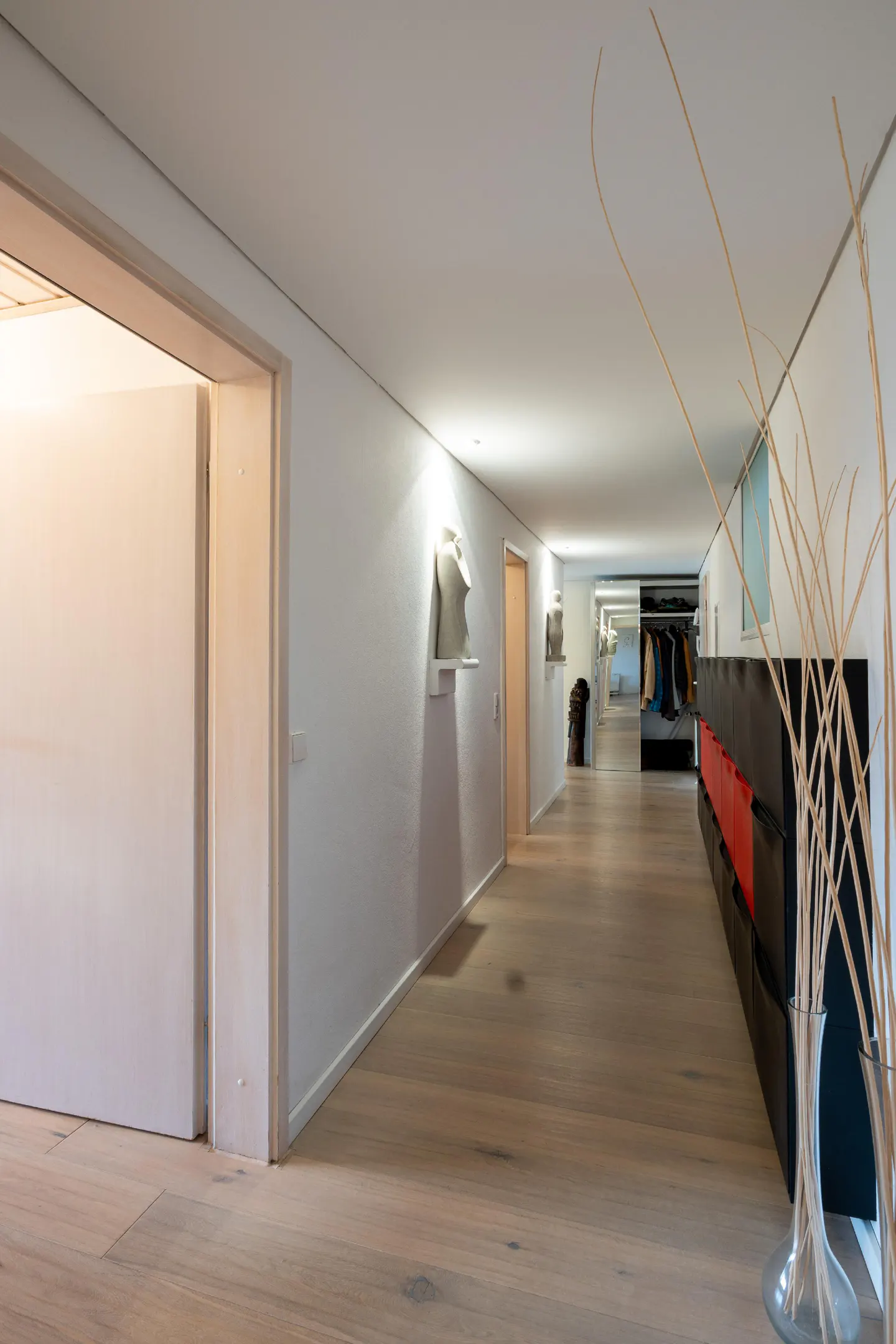 Bright hallway with light wood floors, white walls, and recessed lighting. Sculptures line the wall, and a black storage unit with red bins sits to the right.