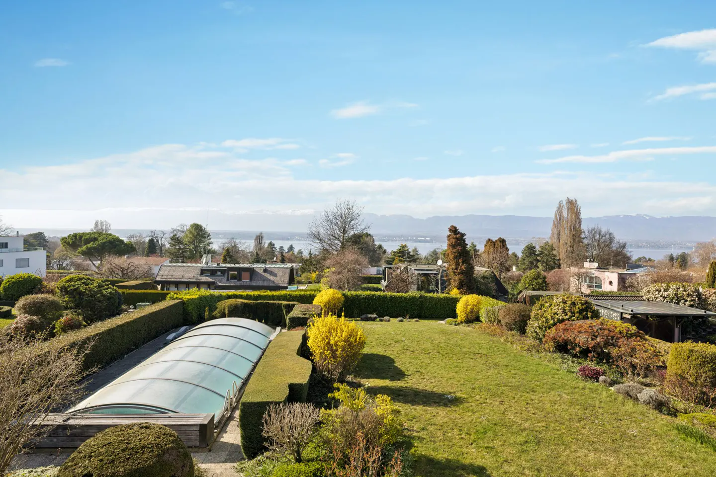 A high angle view of a backyard with a pool, green grass, bushes, and a lake in the background.