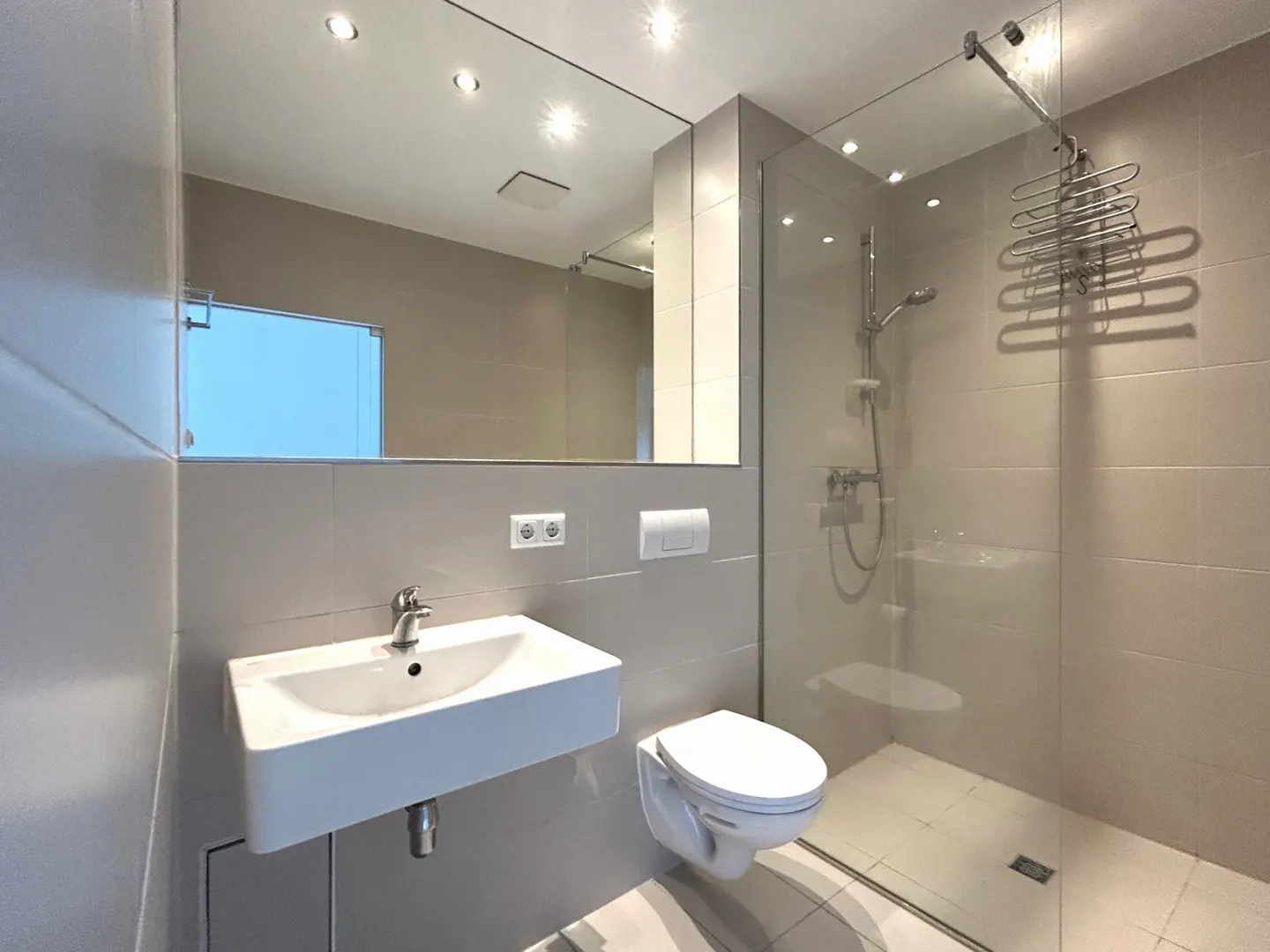 A modern bathroom with gray tiles, a white sink, a toilet, and a glass-enclosed shower with a showerhead and hangers.