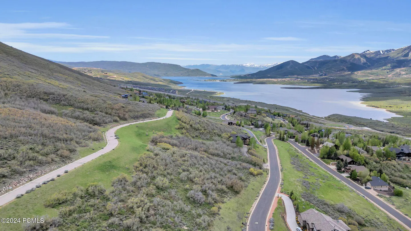 Aerial view of a lake surrounded by green hills and mountains under a blue sky. Roads wind through houses on the hillside.