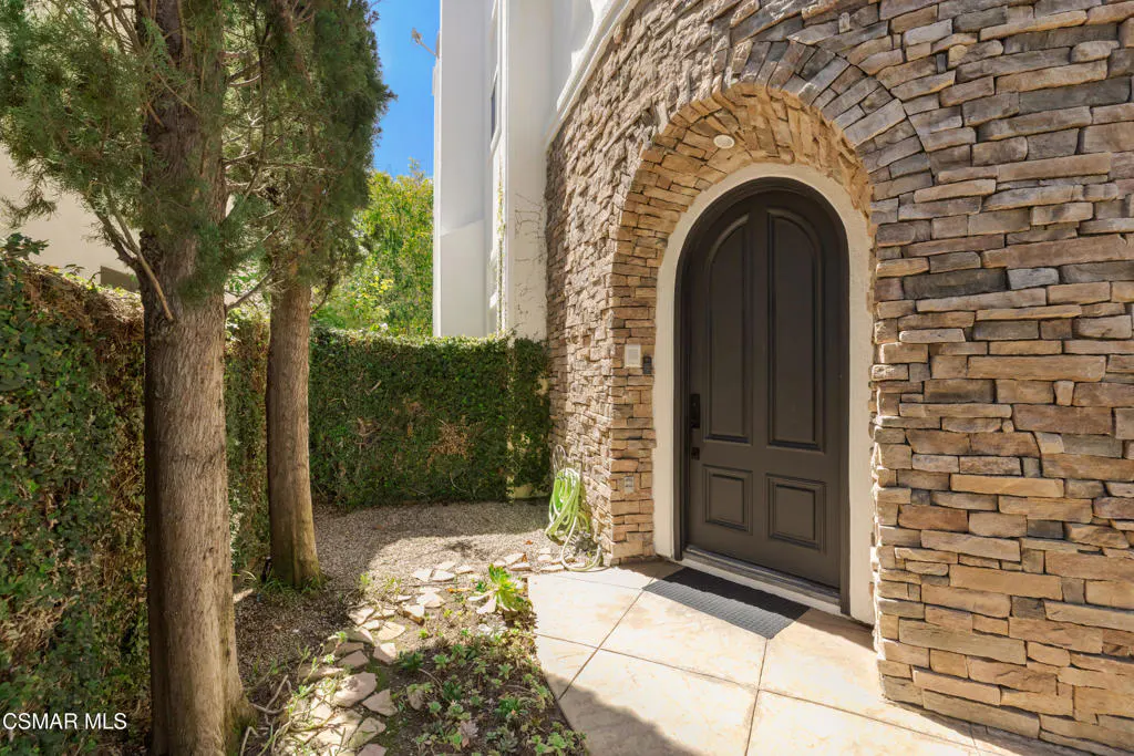 Arched stone doorway with a dark brown door. A stone path leads to the entrance, lined with trees and a green hedge.
