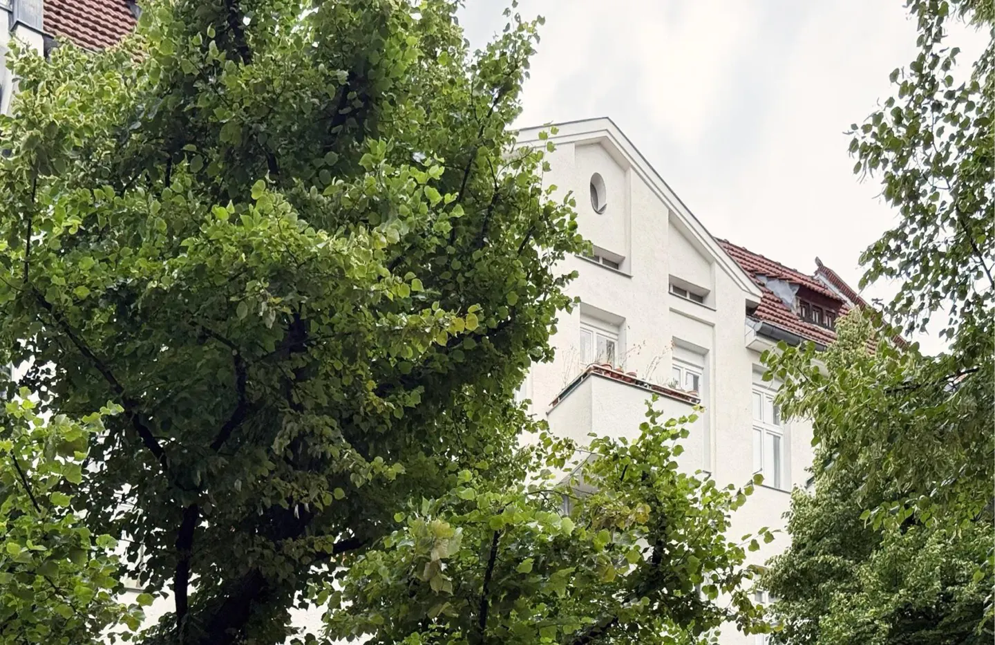 A white apartment building with a red roof is partially obscured by green trees.
