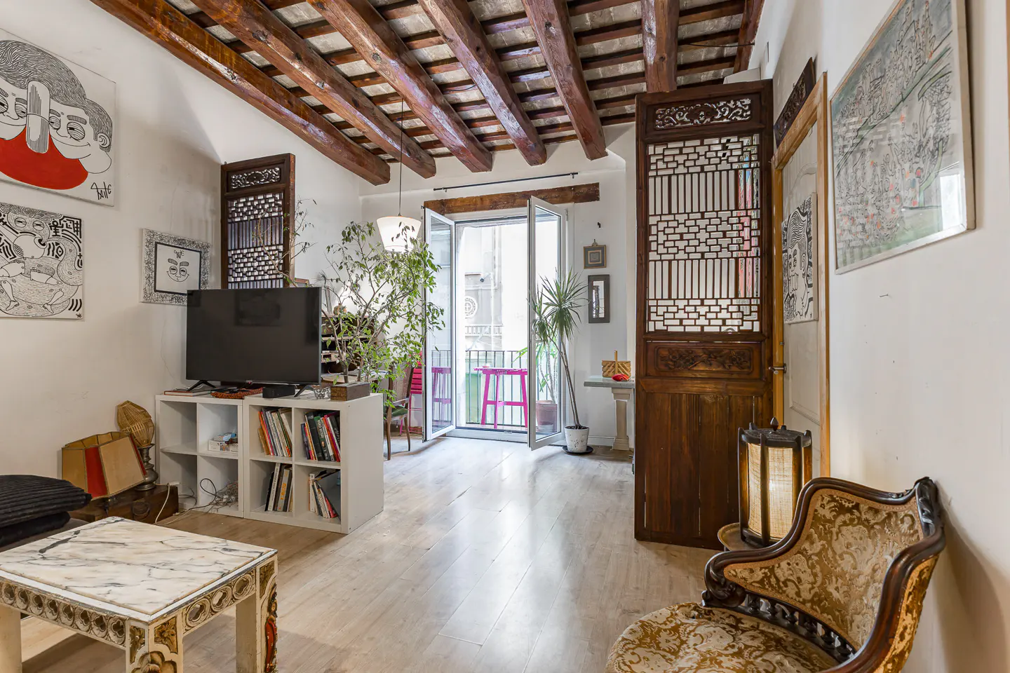 Bright living room with wood-beamed ceiling, white walls, and light wood floors. Balcony with pink table visible through open doors. Art on walls.