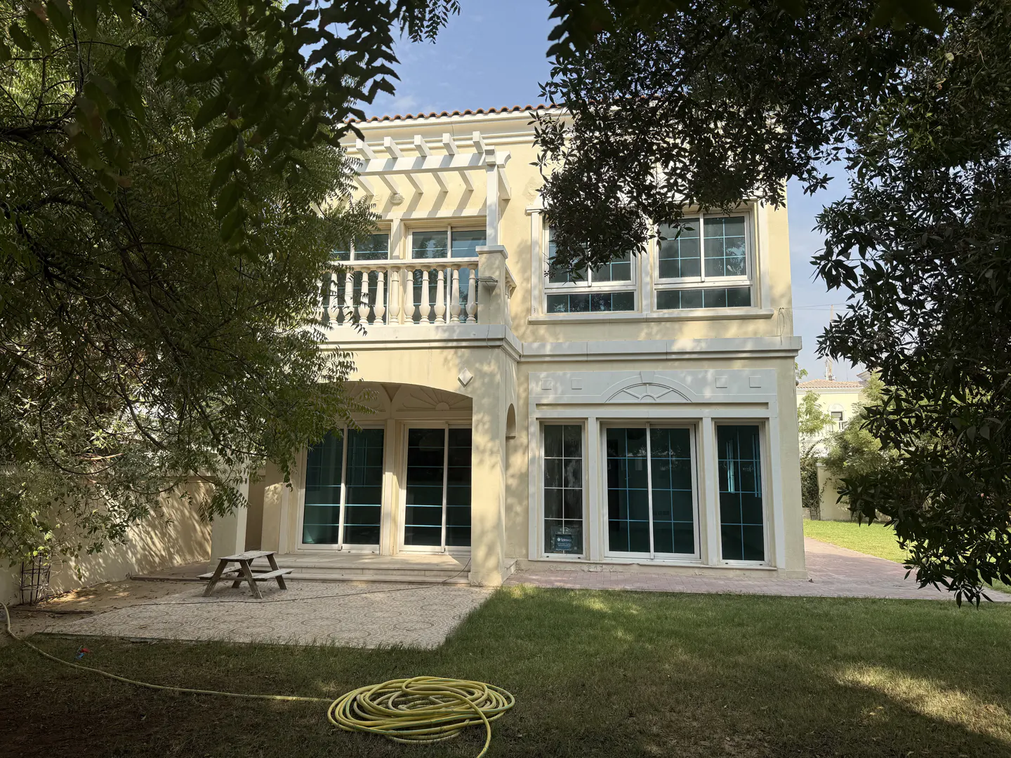 Two-story beige house with a balcony, large windows, and a green lawn. A picnic table sits on a gravel area, and a yellow hose is coiled on the grass.