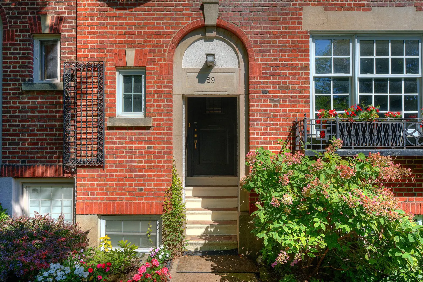 Red brick building with a black door under an arched entrance marked "29". Windows, flowers, and greenery surround the building.