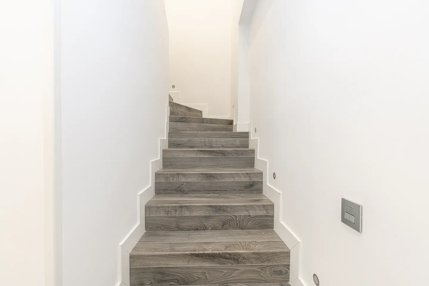 Interior shot of a staircase with gray wood steps and white risers, leading upwards in a white-walled space.