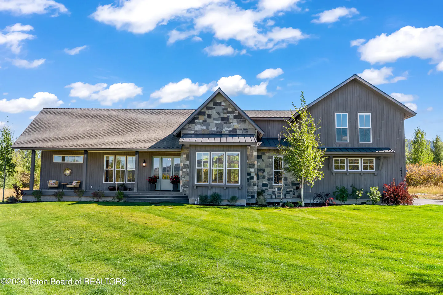 Gray house with stone accents, a porch, and a green lawn under a blue sky with white clouds.