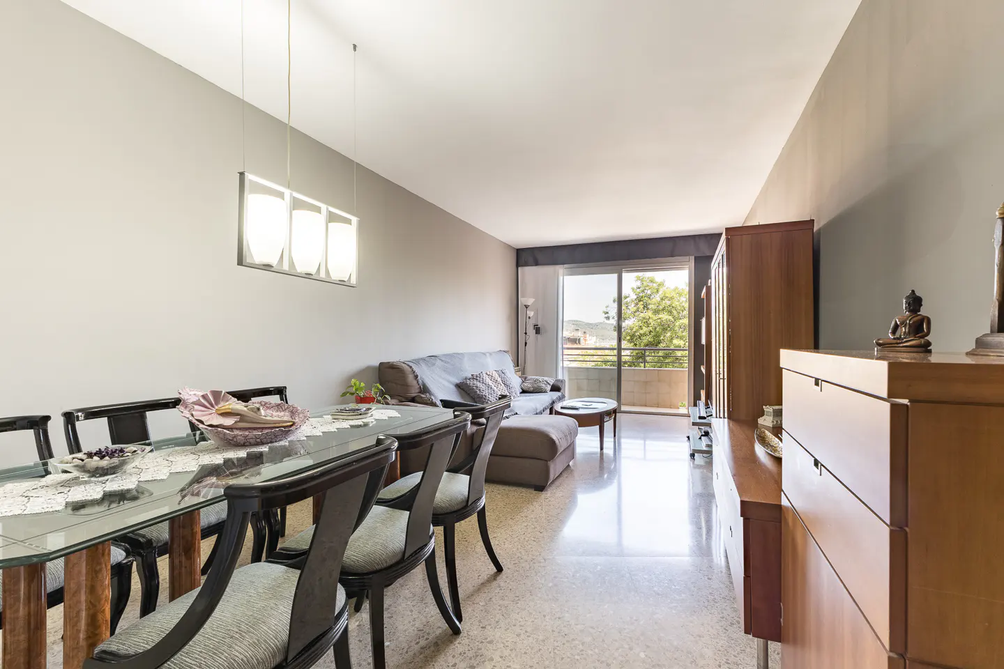 A bright living room with a glass dining table, a gray sofa, and a balcony with a view.
