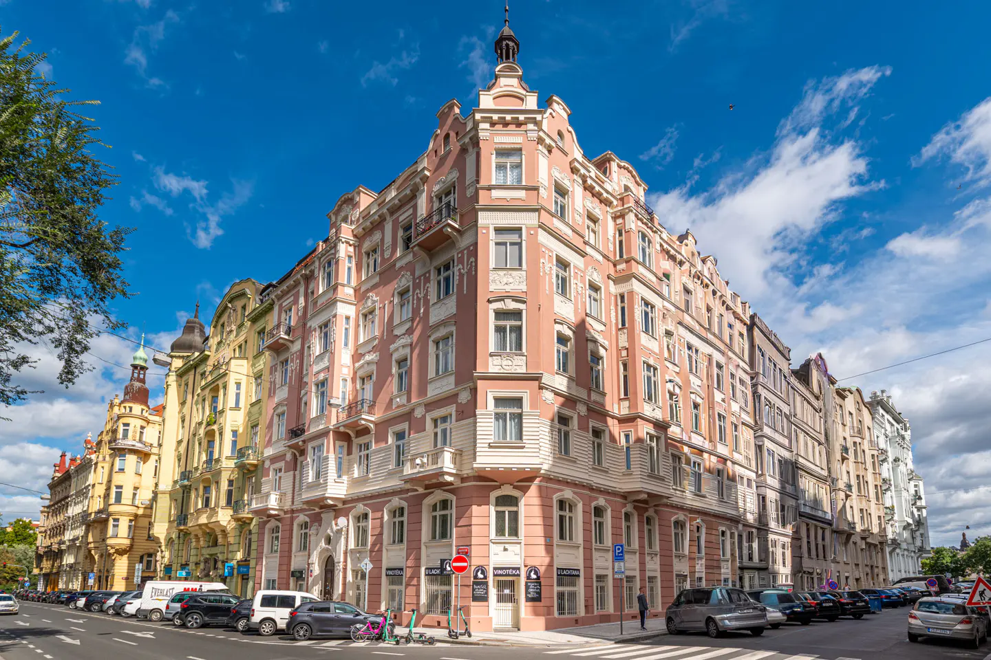 Street view of a pink corner building with white trim, surrounded by other colorful buildings and parked cars under a blue sky.