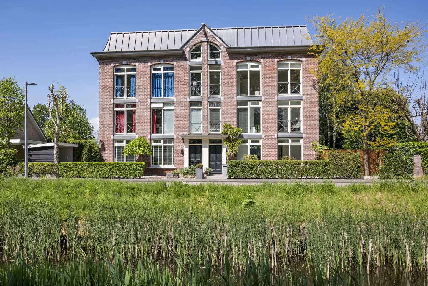 Brick townhouse with a metal roof, white window frames, and colorful curtains, viewed from across a grassy waterway.
