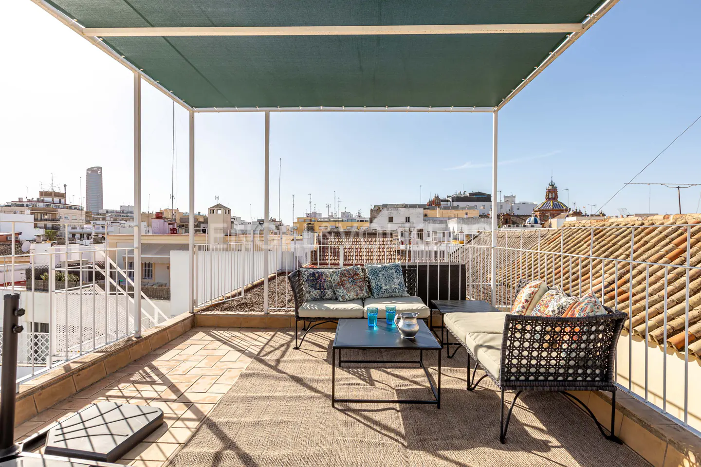 Rooftop patio with wicker furniture, blue glasses, and city views. Green awning provides shade.